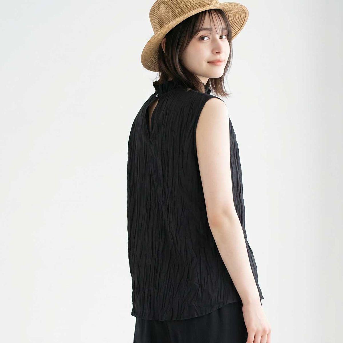 Mikonosublue Storeの黒い綿ローン素材のクリンクル加工ノースリーブブラウス。首元のフリルとバックボタンが特徴的で、麦わら帽子を被った女性が着用している後ろ姿。