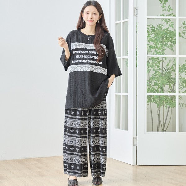 Mikonosublue Storeの胸元にレースとロゴをあしらった黒のビッグシルエット半袖トップスと柄パンツのコーディネート