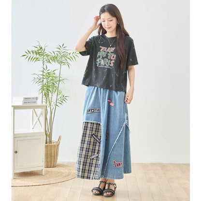 Mikonosublue Storeのデニムとチェック柄を切り替えたパッチワークマキシスカート。ワッペンとフリンジが特徴のリメイク風デザインで、ウエストゴム仕様のコットン製ロングスカートを着用した女性。