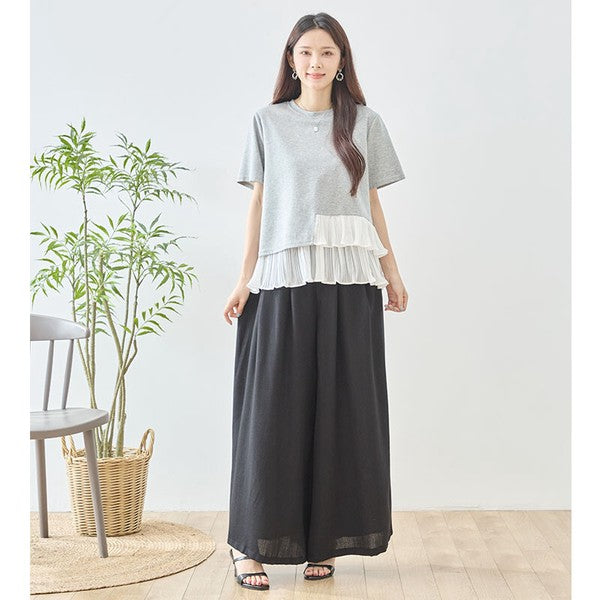 Mikonosublue Storeのグレーの半袖Tシャツ。裾に白いアシンメトリーなプリーツフリルをあしらった異素材切り替えデザインで、モデルが黒のワイドパンツと合わせて着用。