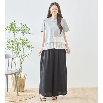 Mikonosublue Storeのグレーの半袖Tシャツ。裾に白いアシンメトリーなプリーツフリルをあしらった異素材切り替えデザインで、モデルが黒のワイドパンツと合わせて着用。