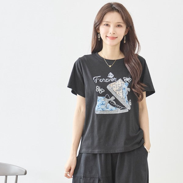Mikonosublue Storeのラインストーンと異素材パッチワークのスニーカー柄が施された黒の半袖Tシャツを着た女性